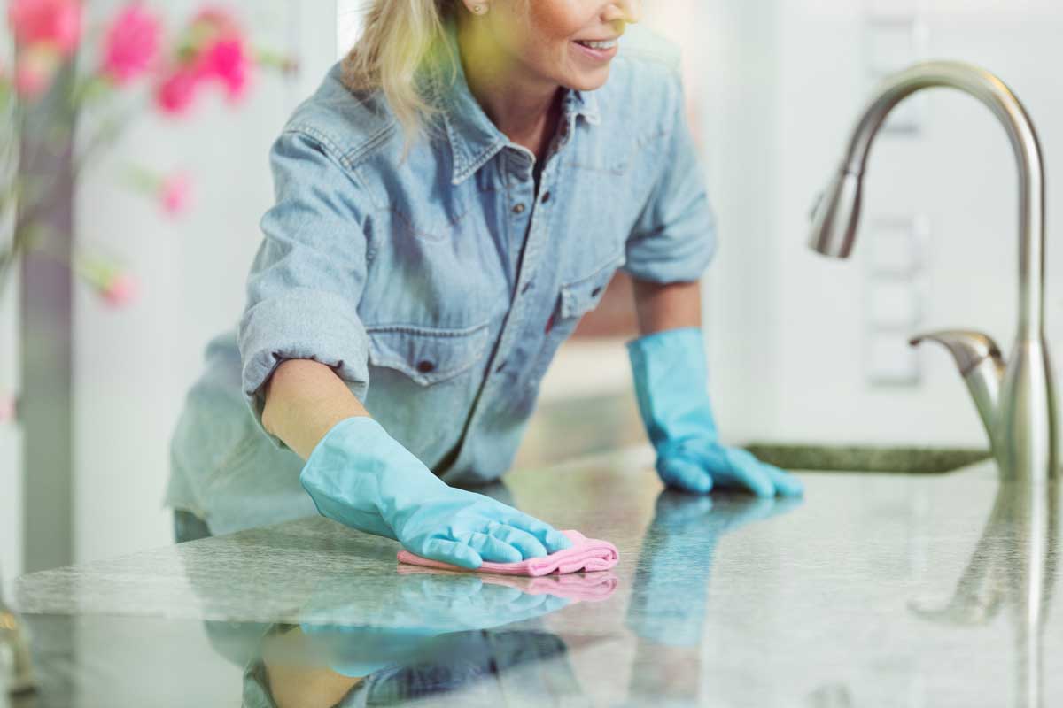 Pedantic woman wiping down kitchen countertop with pink cloth, wearing blue jean shirt and rubber gloves for household cleaning