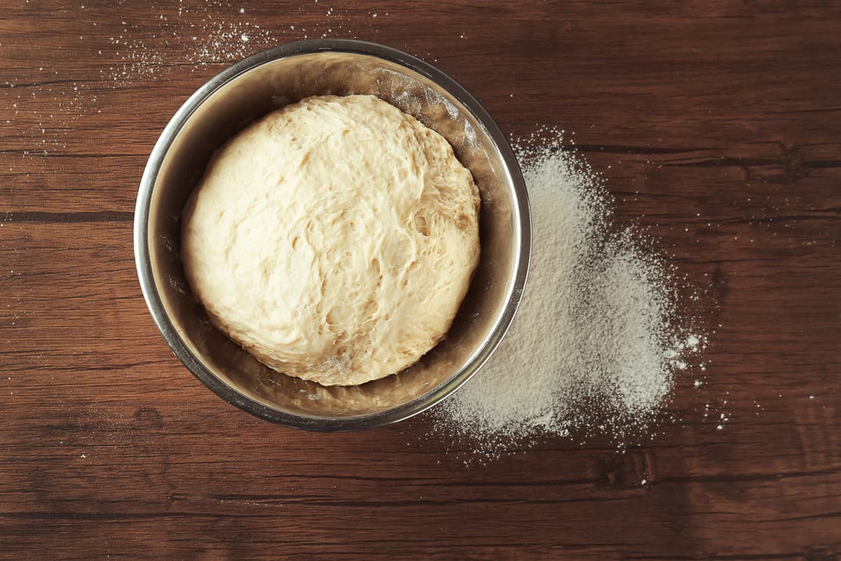 Raw bread dough on a stainless steel mixing bowl with flour spread on a wooden table