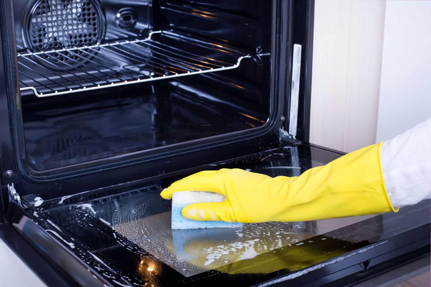 A woman wearing gloves and using a sponge to clean the glass door of an oven
