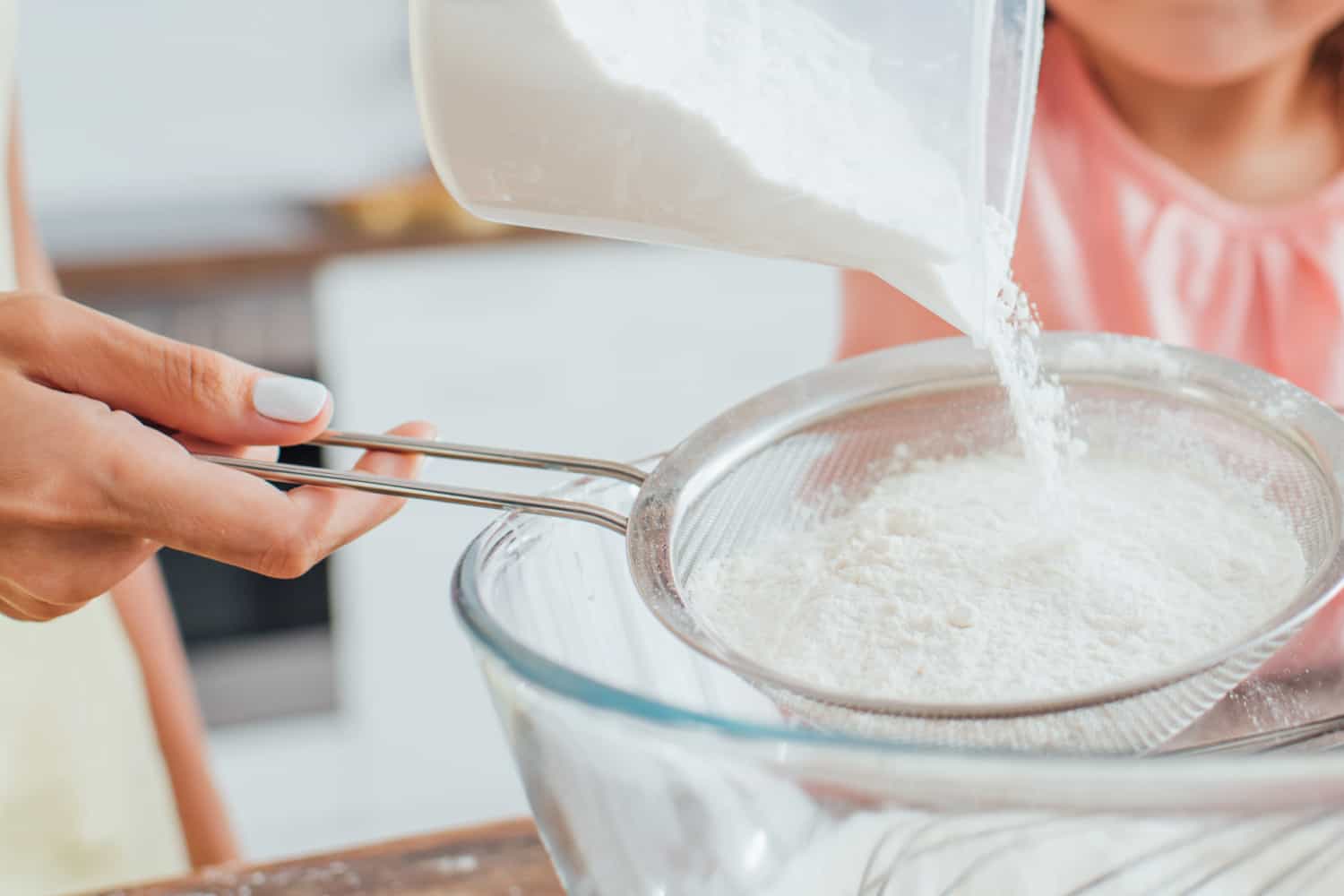 woman sifting flour into a glass bowl with daughter watching