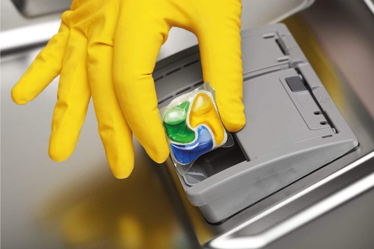 Close up of hand in yellow rubber glove putting detergent tablet into dishwasher