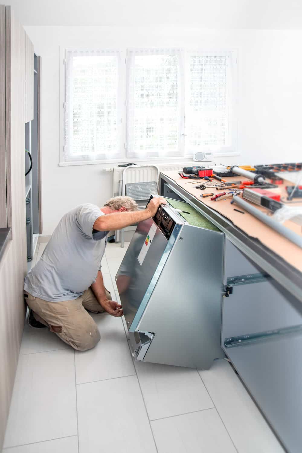 Vertical shot of kitchen fitter technician men, manual worker plumber, mature men gray hair, installing new appliance integrated furniture dishwasher in a new kitchen custom made