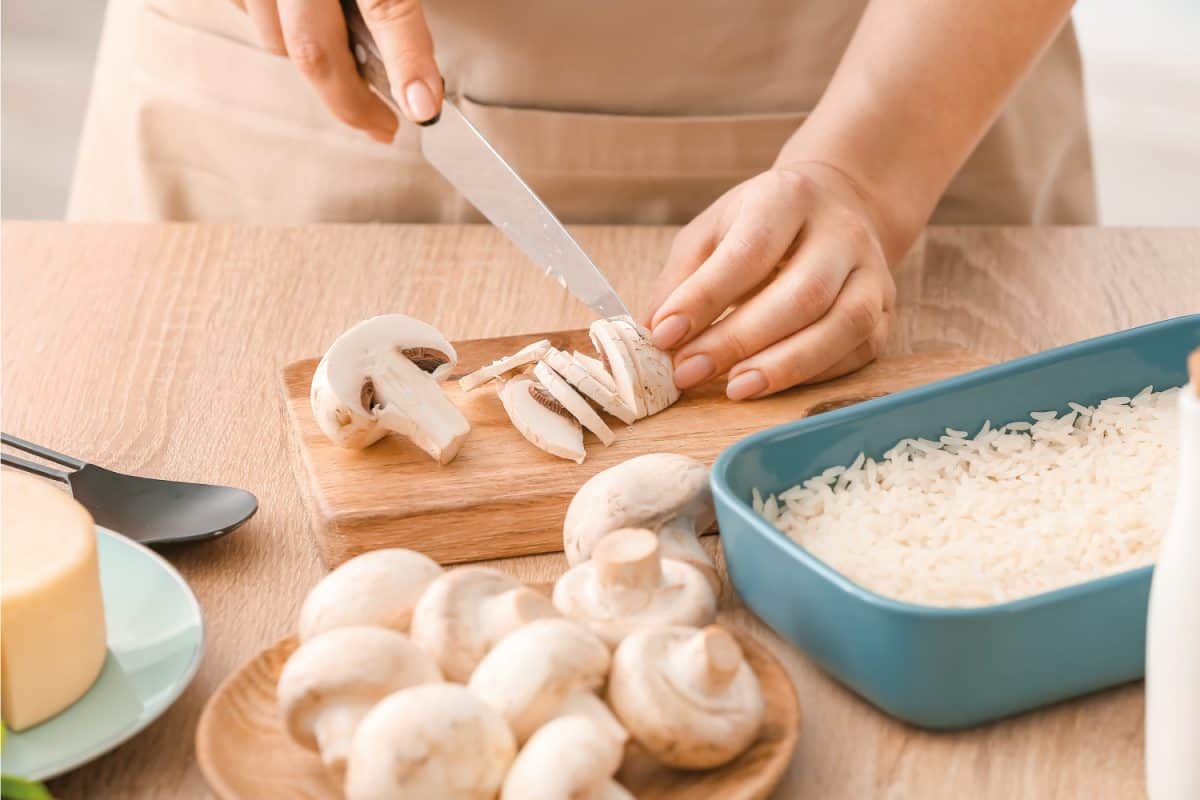 Woman cooking rice casserole on table, chopping mushrooms on chopping board