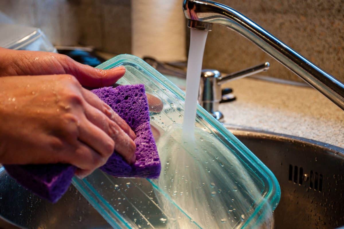 Woman washing a plastic food storage container with a sponge in kitchen sink.