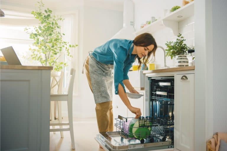 Woman wearing apron loading soiled dishes in a dishwasher in bright kitchen, How To Clean Bosch Dishwasher [5 Steps]