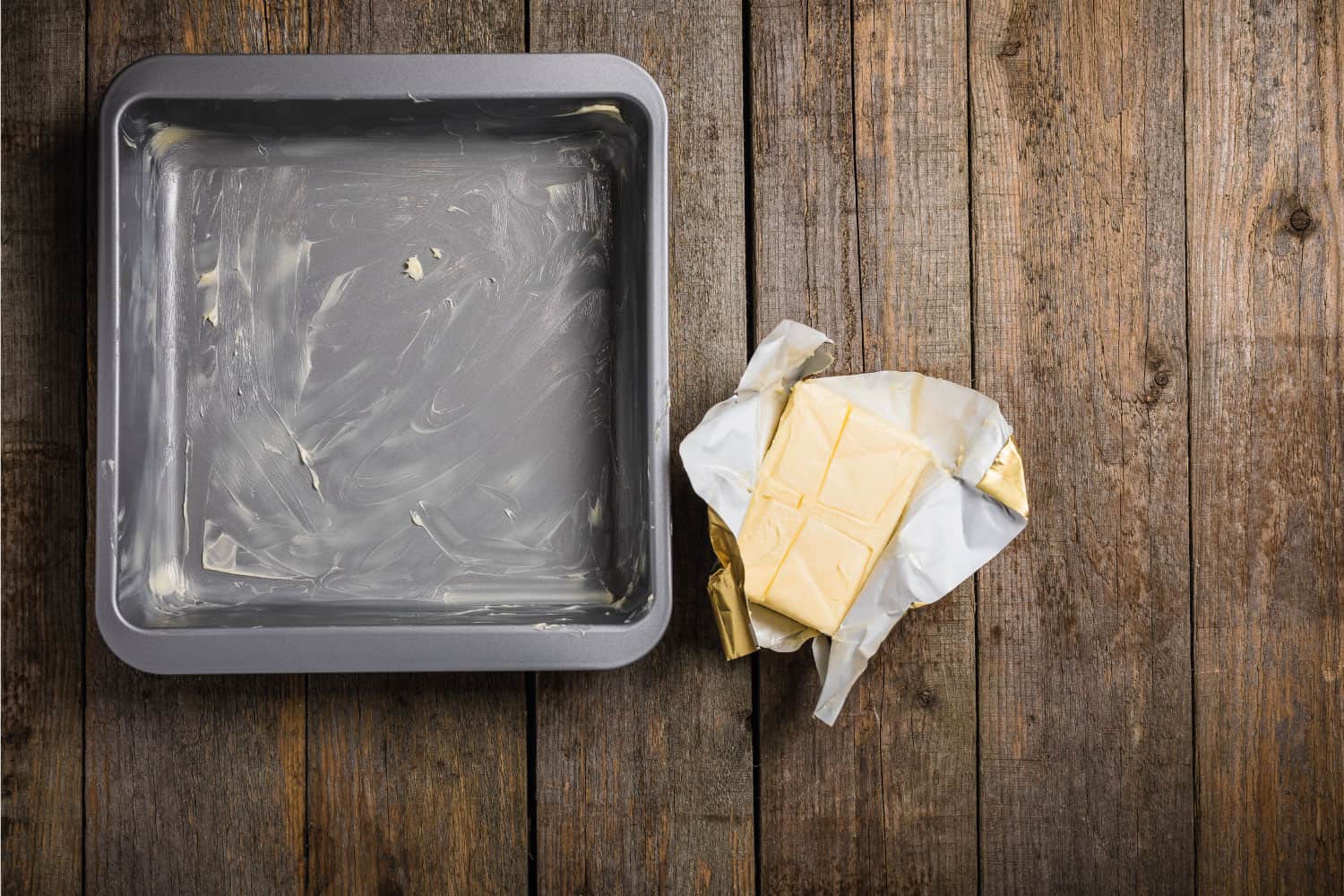Non stick square cake tin, greased with butter and an open packet of butter sat on a rustic wooden surface