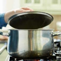 Woman lifting steel lid over cooking pan on stove, Cooking: Covered Vs Uncovered (When Should You Cover A Cooking Pot?)