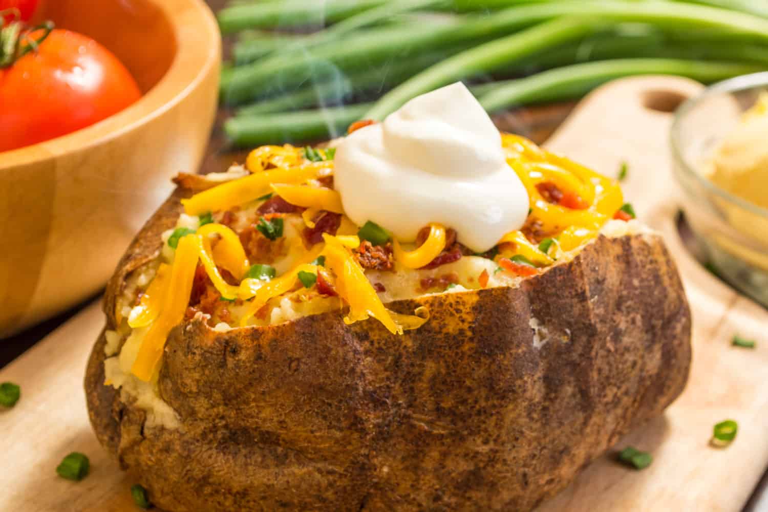 fresh hot baked potato with butter bacon cheese and chives topped with sour cream on a cutting board