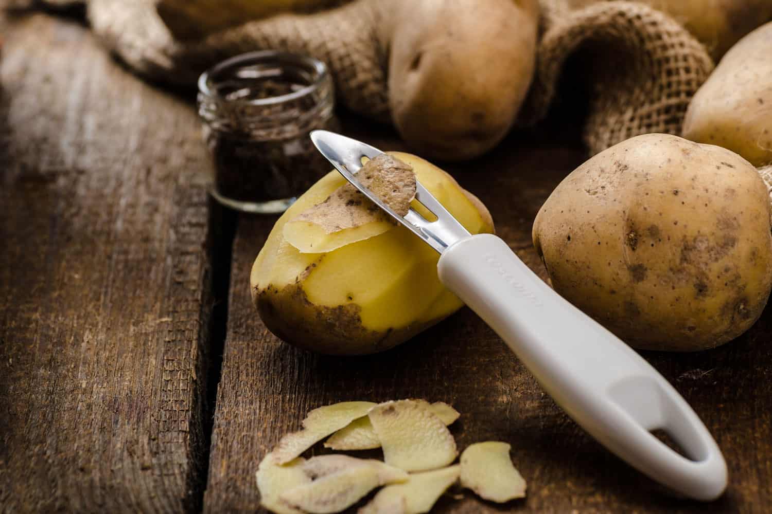A peeled potato with a potato peeler on it and unpeeled potatoes on the background