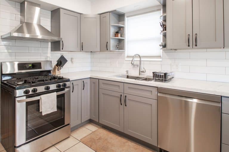 Interior of a modern kitchen with gray colored cabinet panels, a kitchen range with a hood, and a white tiled plank backsplash, How To Protect Kitchen Cabinet Doors From Water Damage