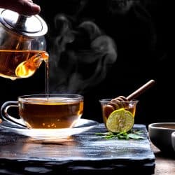 Pouring hot aromatic herbal tea from teapot into glass teacup set with steam and various herbs on black stone plate with wooden table floor in dark background, 5 Best Saucepans For Making Tea