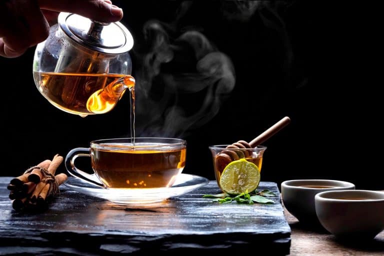 Pouring hot aromatic herbal tea from teapot into glass teacup set with steam and various herbs on black stone plate with wooden table floor in dark background, 5 Best Saucepans For Making Tea