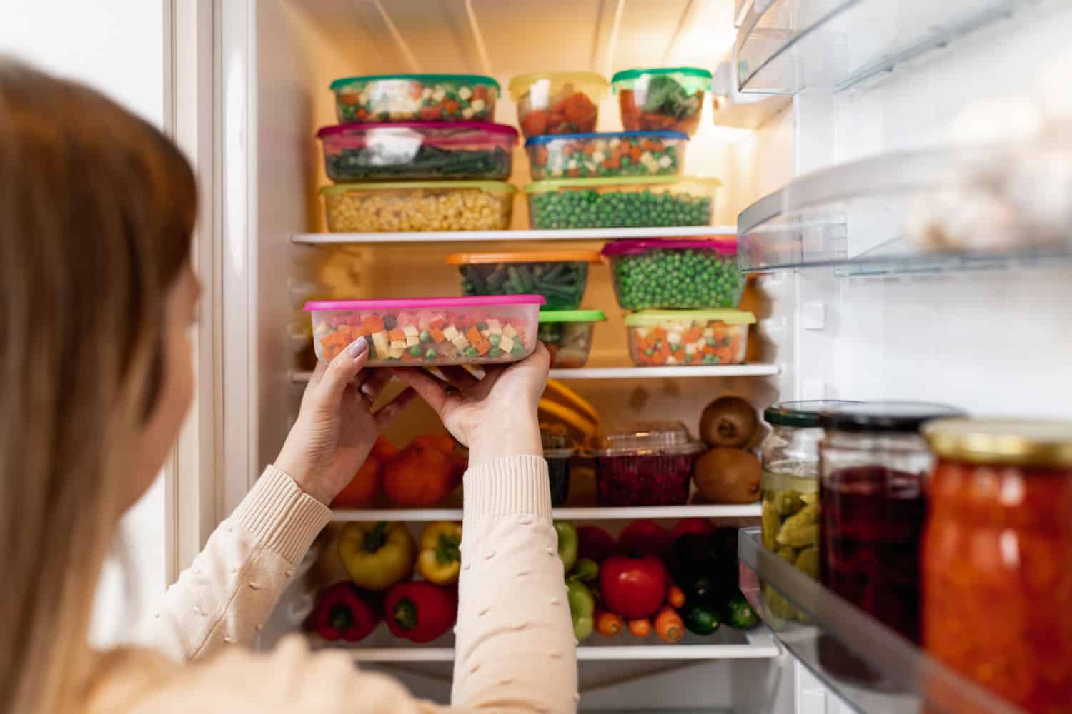 Close up shot of woman taking container with frozen mixed vegetables from refrigerator while looking at camera
