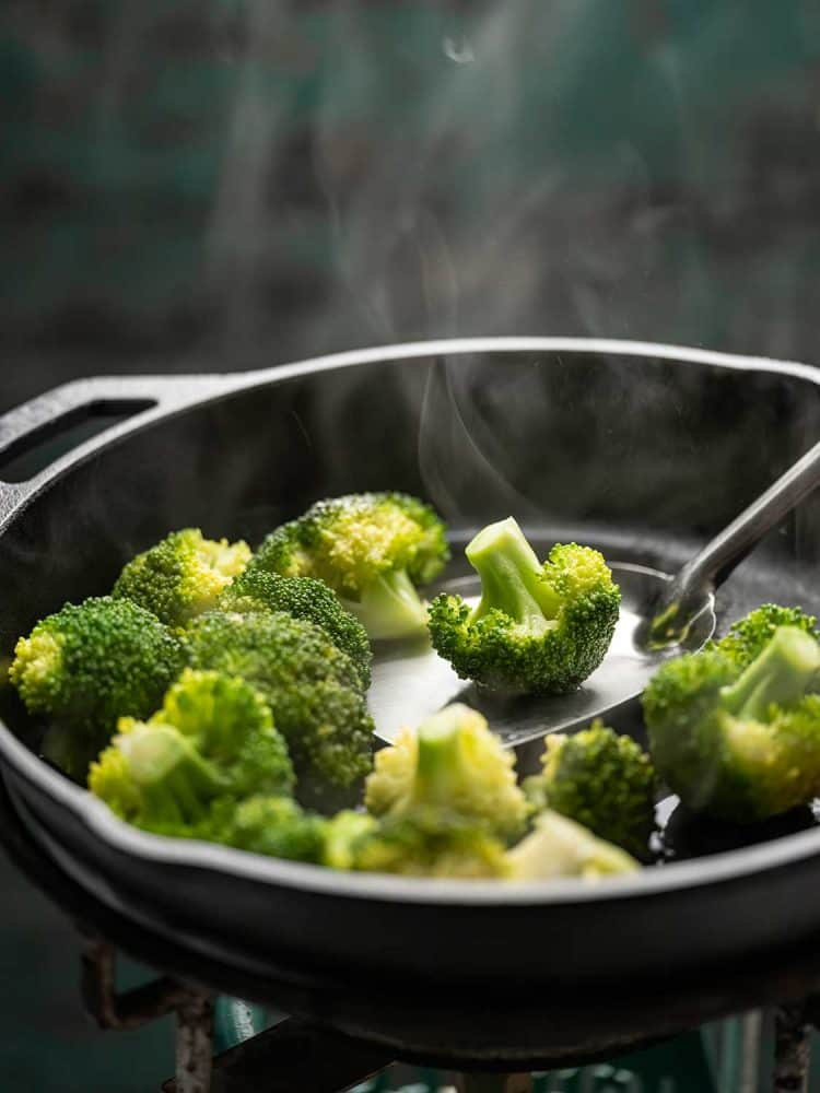 Fresh broccoli spears being stir fried in a heavy iron pan on a gas stove