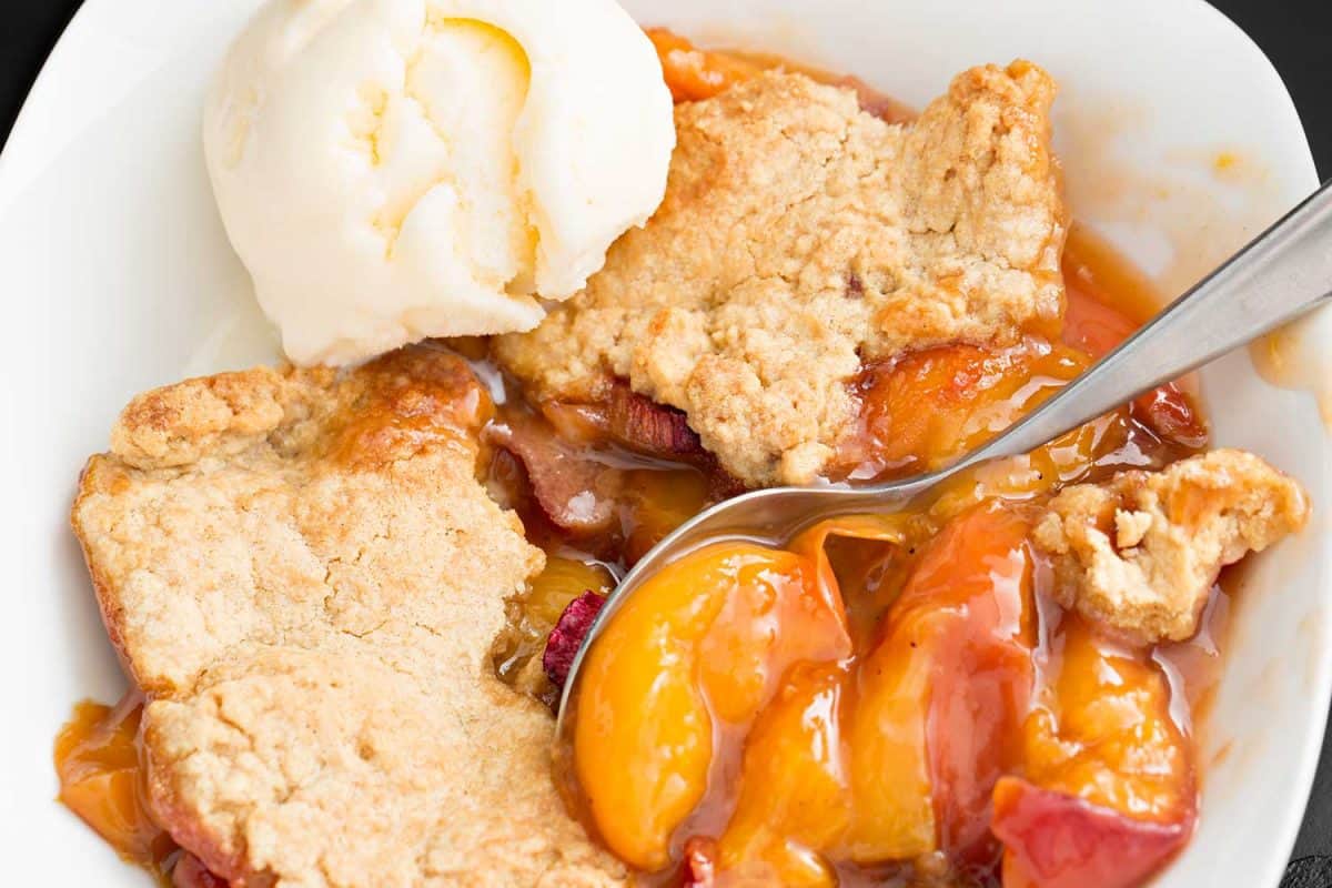 An overhead close up of a peach and rhubarb cobbler with a scoop of vanilla ice cream in a white bowl