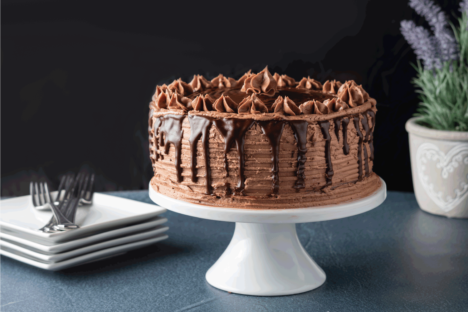Decadent chocolate cake on a white cake platter. Cake is iced with chocolate buttercream frosting and drizzled with a chocolate ganache. White square plates and forks adorn the table