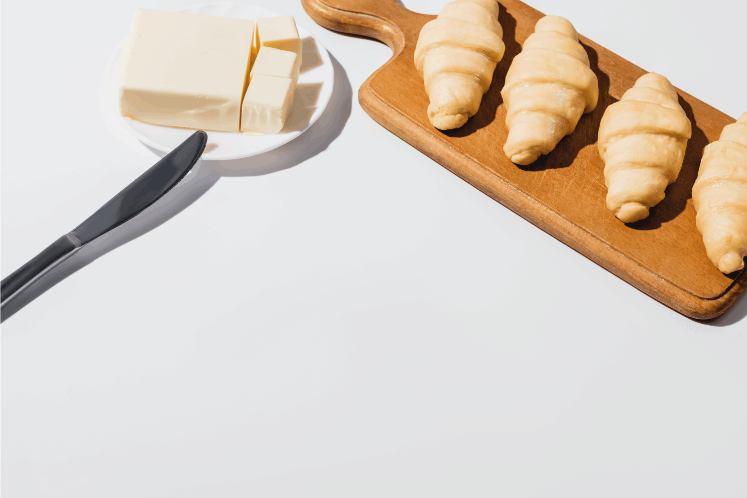 Fresh raw croissants on wooden cutting board near butter on plate with knife