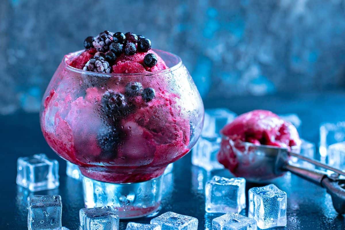 Fruit and berry ice cream in a large beautiful glass decorated with leaves of mint and scoop, surrounded by ice cubes on black stone background