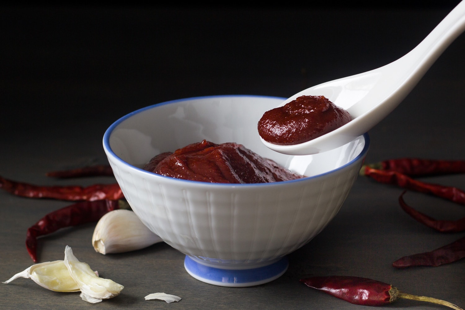 Gochujang in white bowl with spoon, with garlic and chilis on dark background