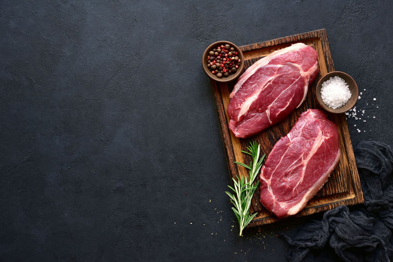 Raw organic marbled beef steaks with spices on a wooden cutting board on a black slate, stone or concrete background, Should You Always Marinate Steak? [Inc. 11 Awesome Steak Marinade Ideas]