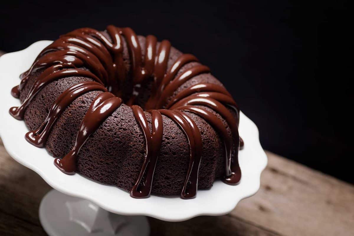 A chocolate bundt cake with ganache icing sits atop a cake stand on a wooden table