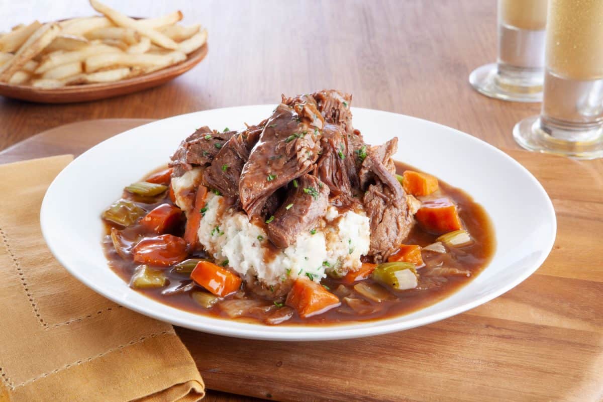 A pot roast placed over mashed potatoes and other vegetation under gravy