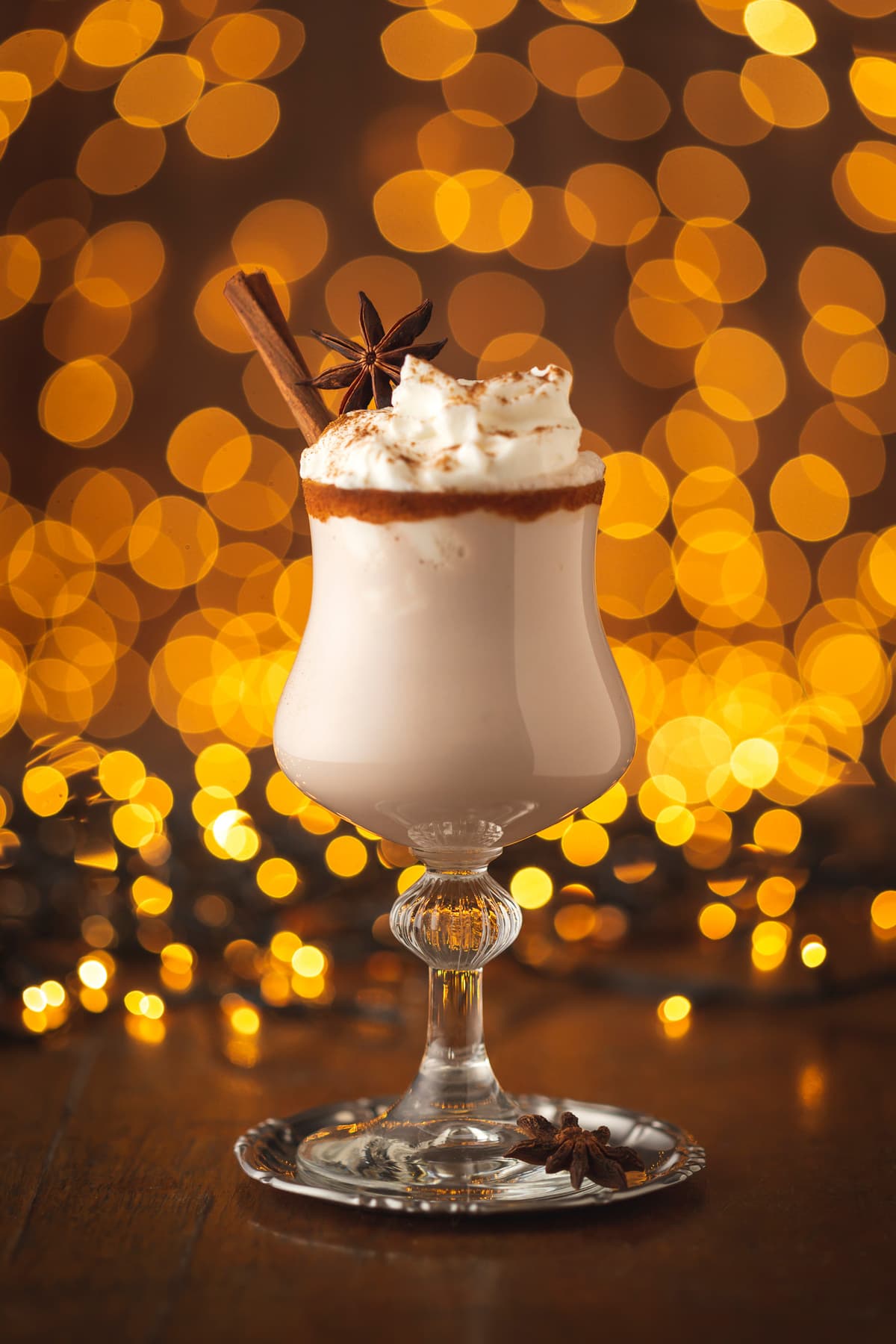 Delicious creamy homemade eggnog served in glasses with cinnamon rim decorated with anise star and cinnamon sticks with Christmas lights in the background