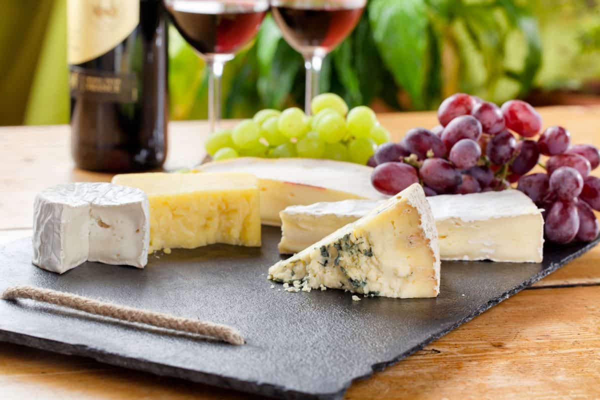 Different slices of cheese on a chopping board with grapes and wine on the side