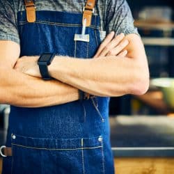 Midsection of confident barista wearing smart watch. Young man is standing with arms crossed. Waiter is wearing apron in coffee shop, 11 Types Of Aprons And Their Uses
