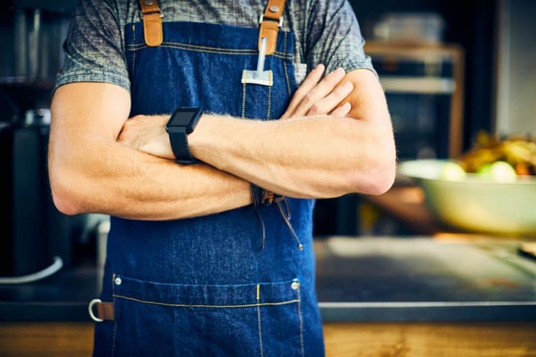 Midsection of confident barista wearing smart watch. Young man is standing with arms crossed. Waiter is wearing apron in coffee shop, 11 Types Of Aprons And Their Uses