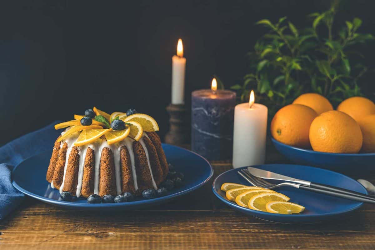 Orange bundt cake with blueberry surrounded fruits, candles, plant and cutleries on dark wooden background