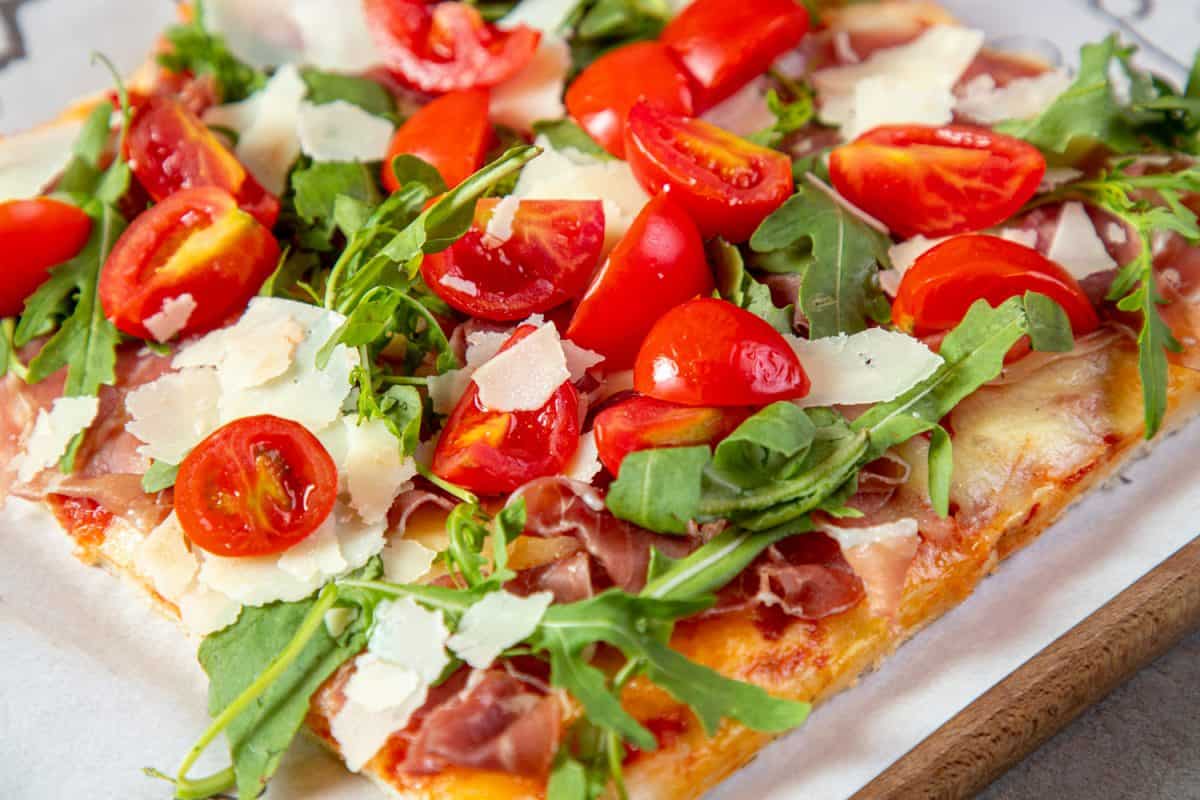Slice of pizza layered with tomato sauce, parmesan cheese and ham, seasoned with arugula, grana cheese and freshly chopped cherry tomatoes