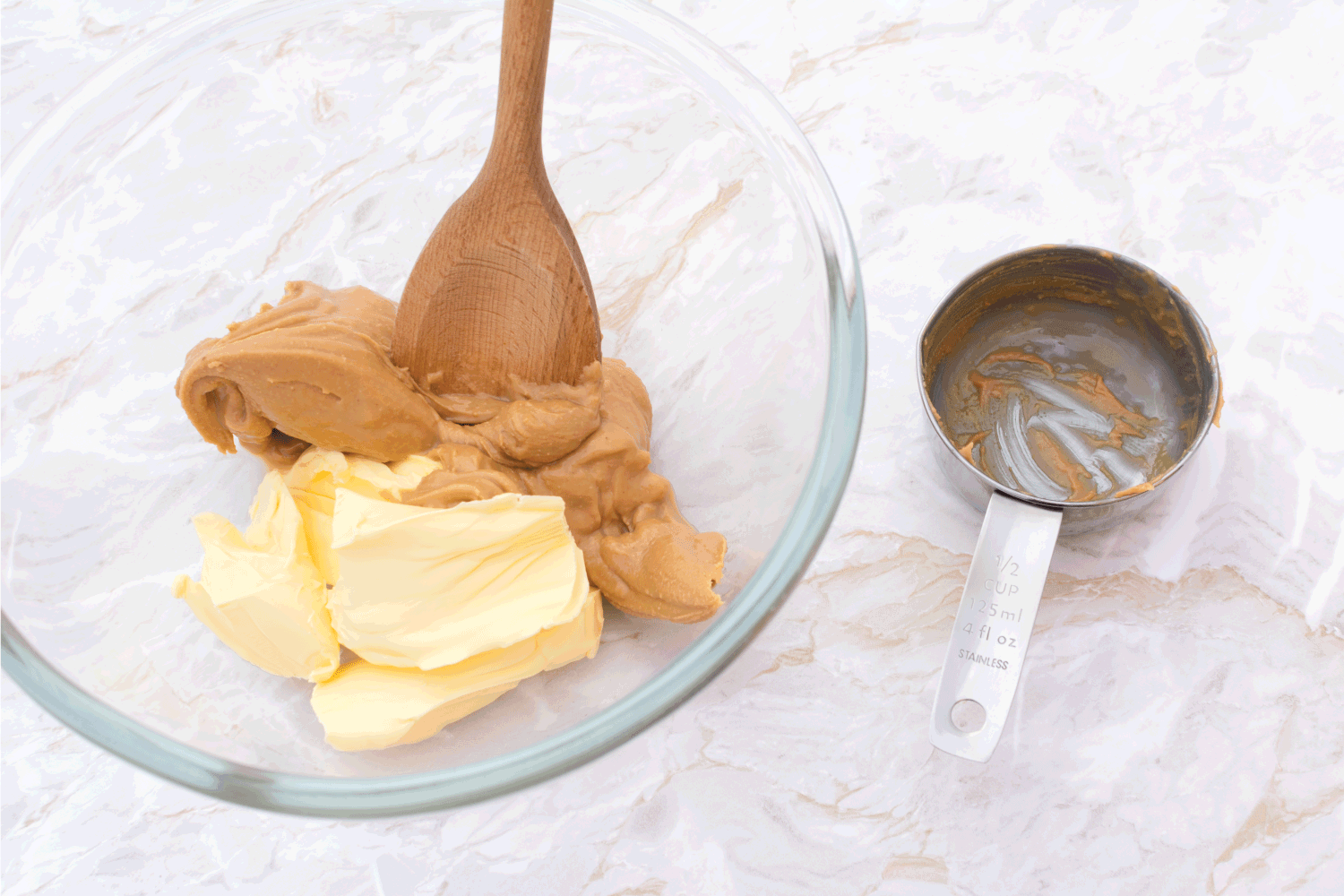 Wooden spoon mixing together smooth peanut butter and butter. Dirty measuring cup sits beside bowl on kitchen counter.