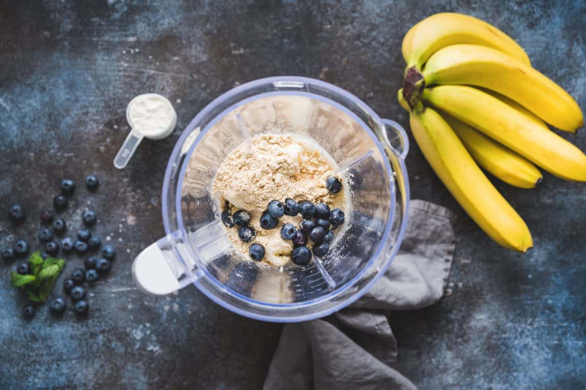 A blender mixed with blueberries, flour, and bananas on the side