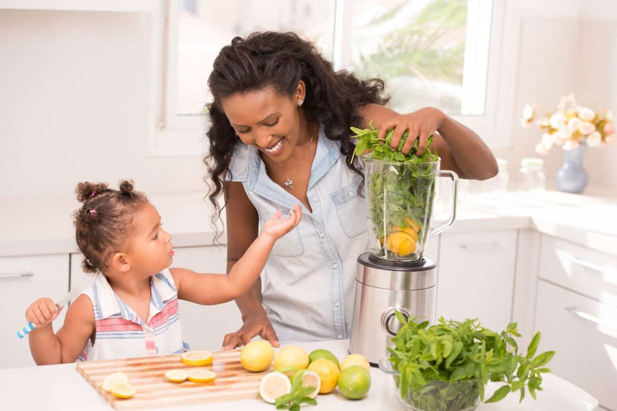 A little girl helping mother at a kitchen. Child cutting lemon-fruit. Mother blending mint leaves