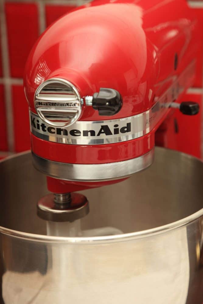 Empire Red coloured Kitchenaid Artisan Stand Mixer, angled front view on red tiled background with flat beater attachment and bowl