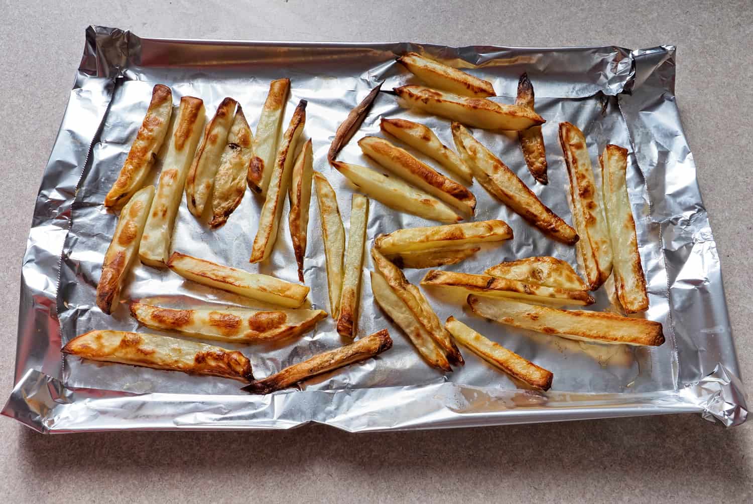 Home prepared baked french fries, in freshly sliced potato and cooked state with olive oil, on a sheet of aluminum foil.