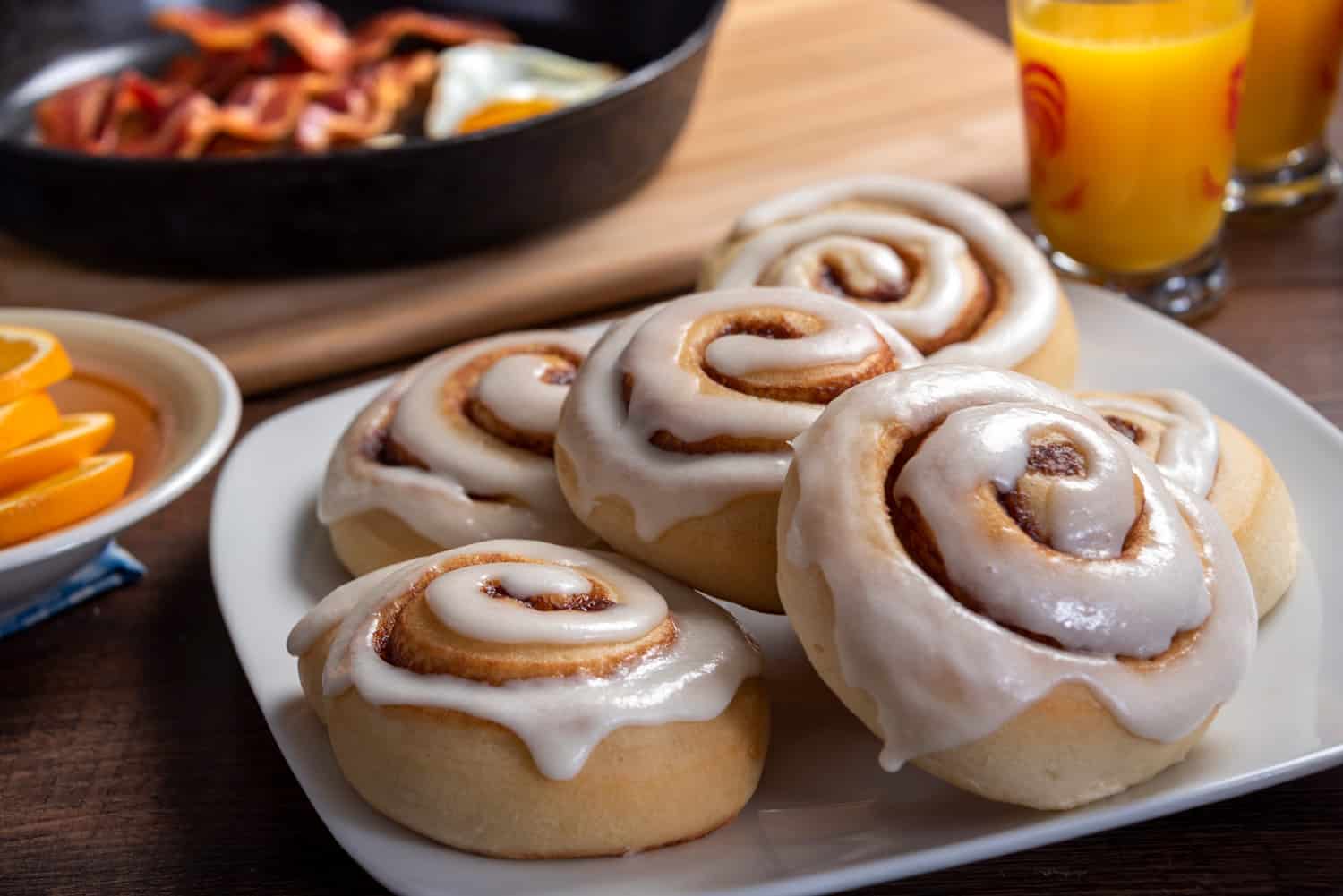 Horizontal photograph of a plate of cinnamon rolls on a breakfast table. Other breakfast items are on the table, but focus is on the plate of large sweet rolls.
