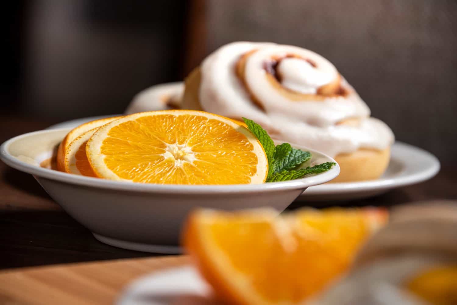 Small bowl of orange slices with a mint garnish sitting on wooden table. Focus is on the orange slices. There is a large cinnamon roll and an orange wedge in the background and foreground out of focus.