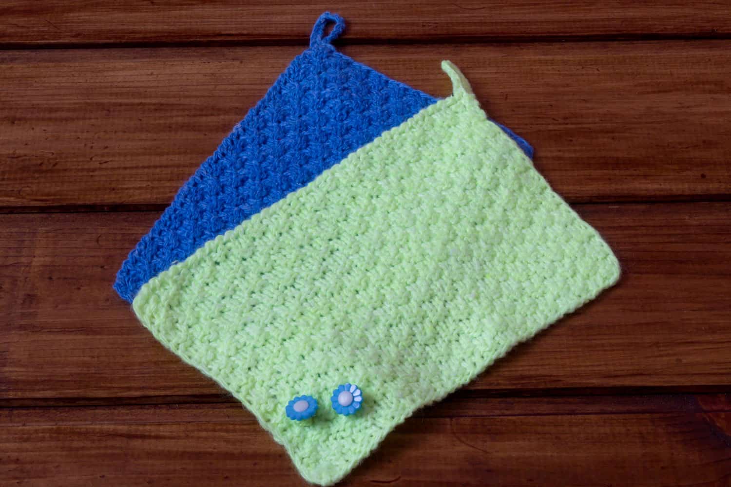 Two hand-knitted napkins or pot holders, green and blue, on wooden background.
