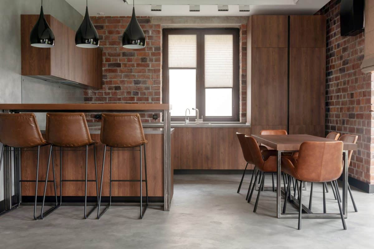 Contemporary interior design of light spacious dinning room including brown wooden furniture with bar stools at counter and soft comfortable chairs at table with cement effect in loft style