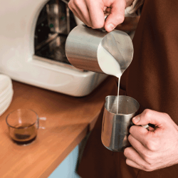 barista-in-brown-apron-pouring-milk-in-steel-jug