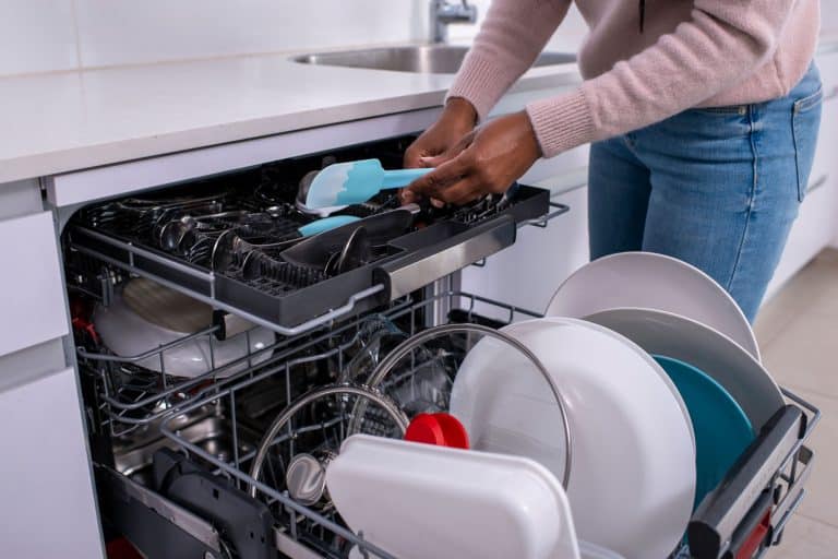 Close up image of a woman's hands holding a spatula, taking out clean cutlery from the dishwasher rack, Do Dishwashers Waste Or Save Water And Electricity?