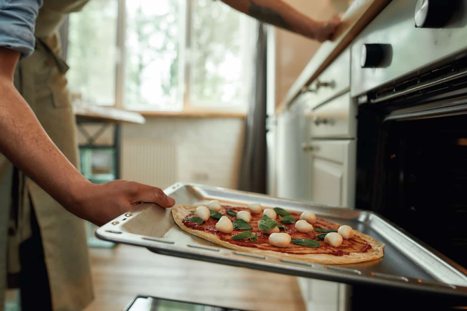 Cropped shot of man, professional cook making pizza at home. Man in apron putting raw pizza in modern oven for baking