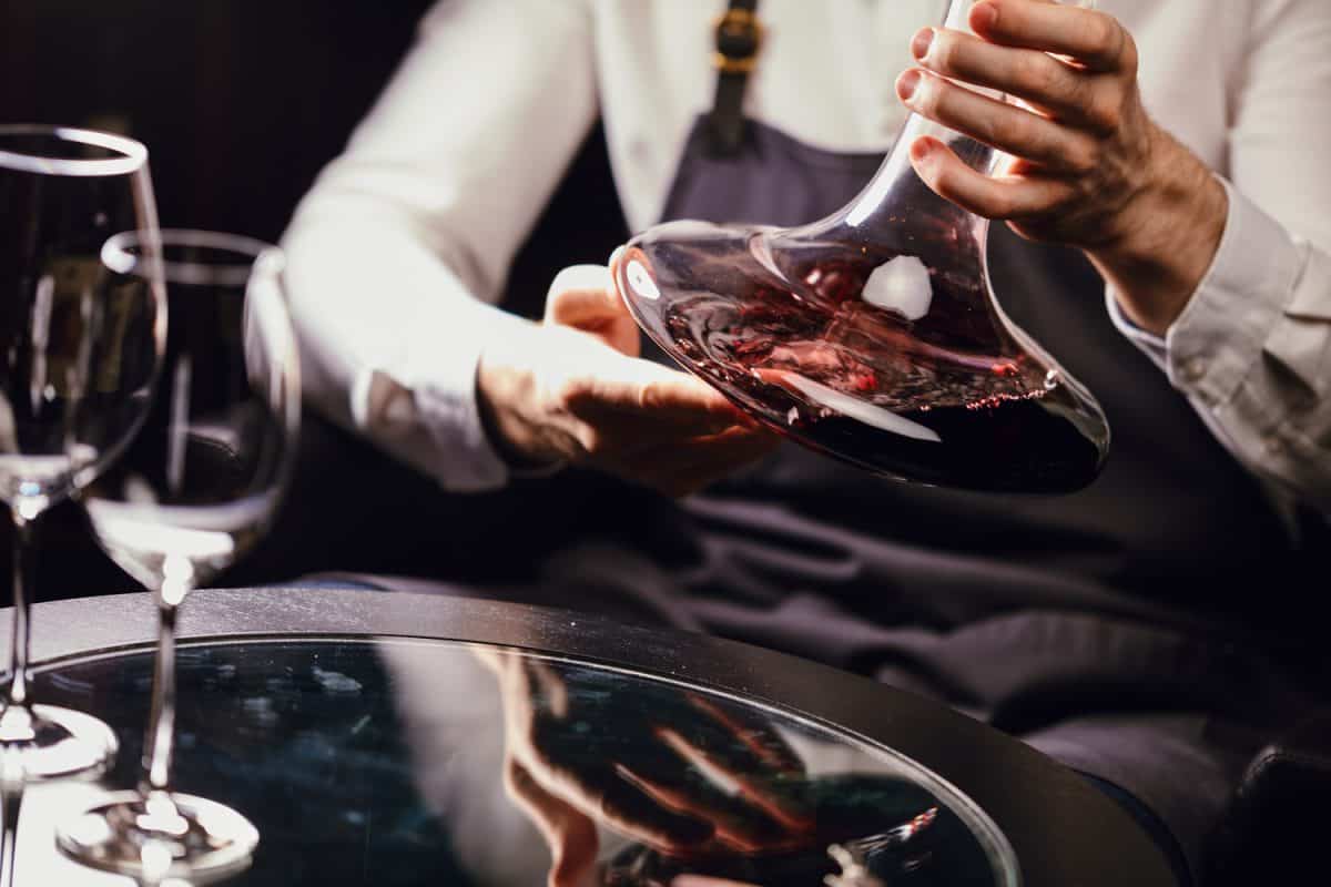 Cropped view of professional sommelier holding mixing bowl with red wine in hands over the table in wine restaurant.