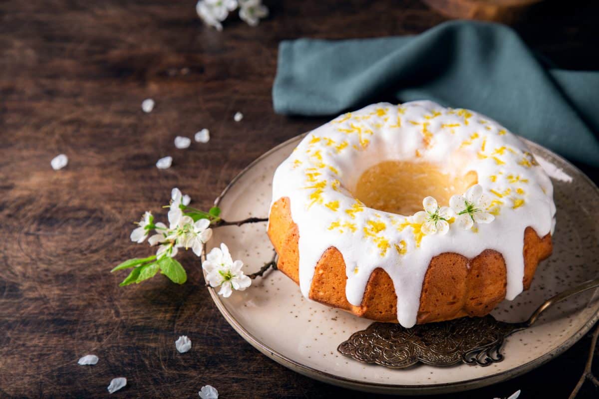 Fresh homemade lemon bundt cake decorated with white glaze and zest on rustic wooden background with branches of blossoming plum