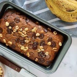 Stock photo showing elevated view of cake loaf tin containing homemade banana loaf, besides a hand of bananas, bowls of chocolate chips and hazel nuts, a tea towel and knife, on a marble effect background., How Full Should A Loaf Pan Be?
