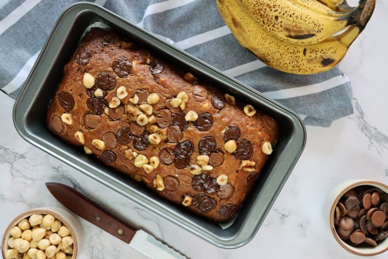 Stock photo showing elevated view of cake loaf tin containing homemade banana loaf, besides a hand of bananas, bowls of chocolate chips and hazel nuts, a tea towel and knife, on a marble effect background., How Full Should A Loaf Pan Be?