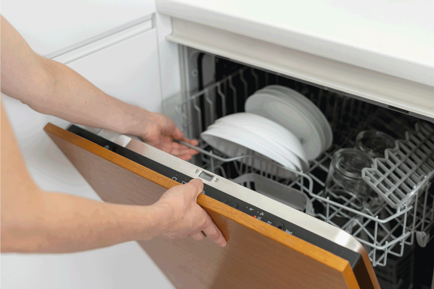Woman opening modern dishwasher machine with plates and cups, standing on kitchen