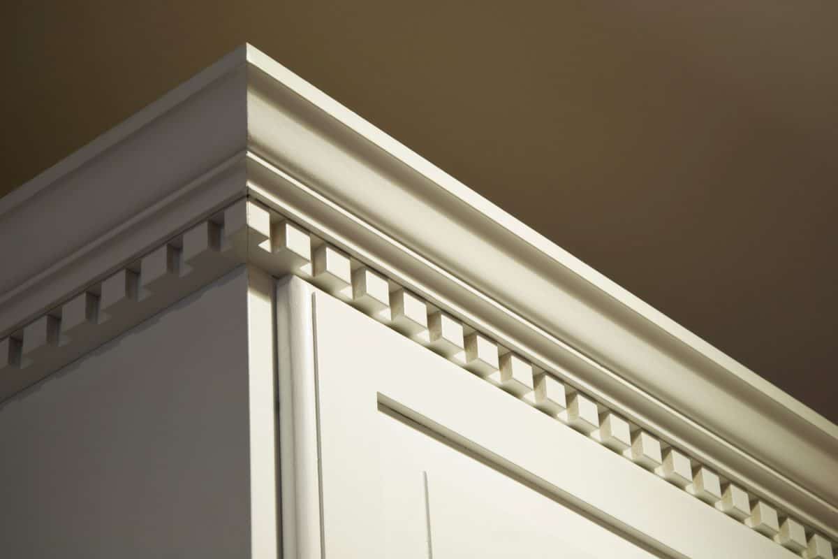 The top crown moulding dentil detail on some creme-color painted solid wood kitchen cabinets. These wall mounted cabinets do not go all the way to the ceiling above them